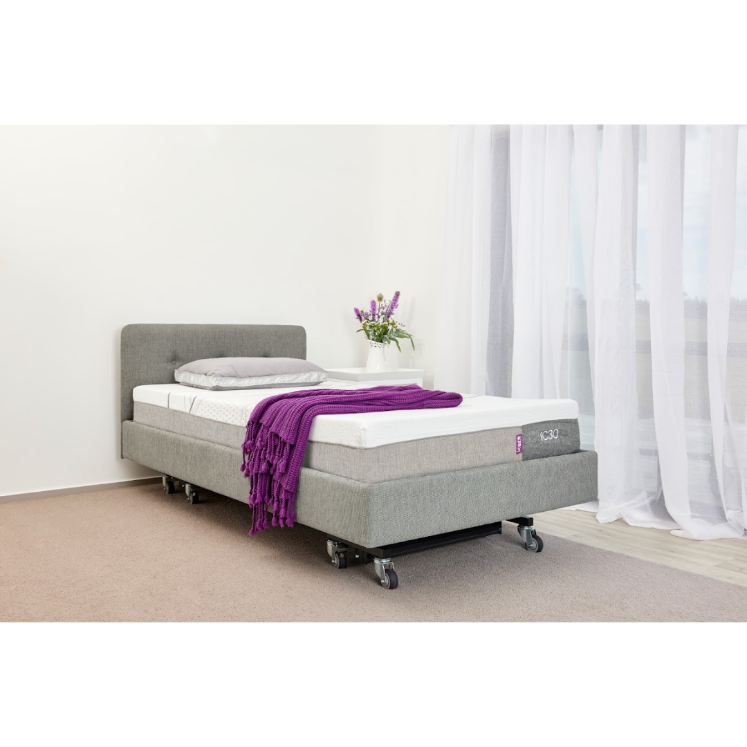 The iCare Medical IC100 Static Partner Non-Adjustable Bed in gray features iCare bed rails and wheels, with a white mattress, gray pillow, and purple blanket. A small white table with flowers stands beside it in a bright room with sheer curtains.